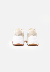 Sportschuhe mit einem beigen strukturierten Obermaterial, weißer klobiger Sohle und braunem Gummi-Außensohle. Verfügt über Zuglaschen an der Ferse für ein einfaches An- und Ausziehen.