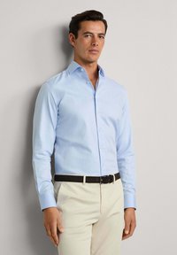 Muškarac stoji uz običan zid, odjeven u svijetloplavu košulju s dugim rukavima, bež hlače i crni remen, gleda naprijed.