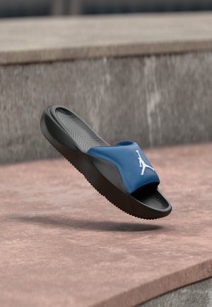 Blau-schwarze Sport-Slide-Sandale mit weißem Jumpman-Logo, das über einer rosafarbenen Steinoberfläche schwebt.