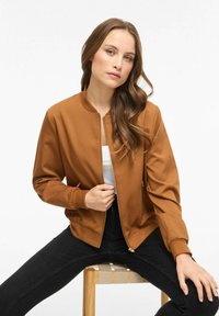 Geacă bomber maro cu fermoar în față, cu manșete tricotate și fermoare la buzunare, purtată peste un top alb și blugi negri.