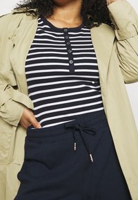 Gestreept marineblauw en wit overhemd met knopen, gecombineerd met marineblauwe joggingbroek met een trekkoord. Bovenlaag is een lichtbeige trenchcoat.