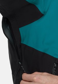 Sort og teal jakke med en glat tekstur. Har et kontrasterende design, lynlås-lomme og en tynd, reflekterende trækflig på lynlåsen.