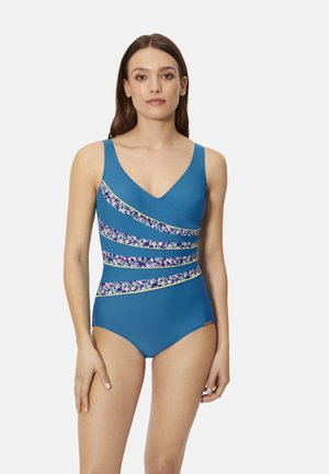 Mujer con un traje de baño de una pieza azul con rayas diagonales florales y ribete amarillo, de pie sobre un fondo claro y liso.