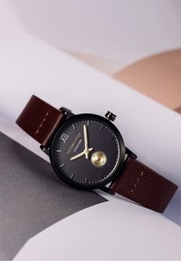Montre noire avec aiguilles dorées et bracelet en cuir marron reposant sur une surface blanche et grise.