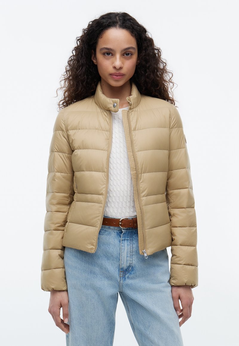 Femme aux cheveux bouclés portant une veste matelassée beige, un pull blanc texturé, une ceinture marron et un jean bleu clair sur un fond uni.