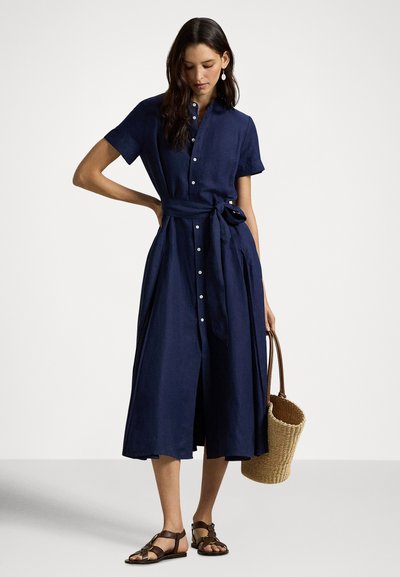 Nježna plava haljina od lana s kratkim rukavima, gumbima sprijeda, remenom za vezanje u struku i lepršavom suknjom, uparena s smeđim sandalama i pletenom torbicom.