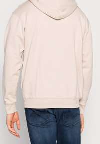 Persona che indossa un hoodie beige chiaro e jeans blu scuro, mostrata di spalle su uno sfondo semplice.