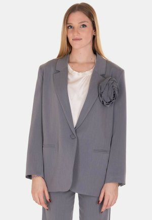 Blazer grigio su misura con rever a scatola, chiusura a un bottone, tasche frontali e un'accenzione in tessuto a forma di rosa sul lato sinistro. Blusa di seta sottostante.