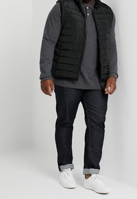 Czarna pikowana kamizelka na szarym, długim rękawie polo, zestawiona z ciemnymi dżinsami i białymi trampkami. Kamizelka ma zamek błyskawiczny i pikowany wzór.