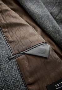Veste en chevrons grise ouverte révélant la doublure intérieure marron à motifs et la poche intérieure bordée de noir avec une étiquette sur le bord inférieur.