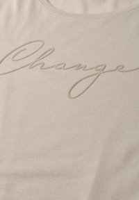 Light beige fabric with raised cursive text spelling "Change" across the chest area of a shirt.