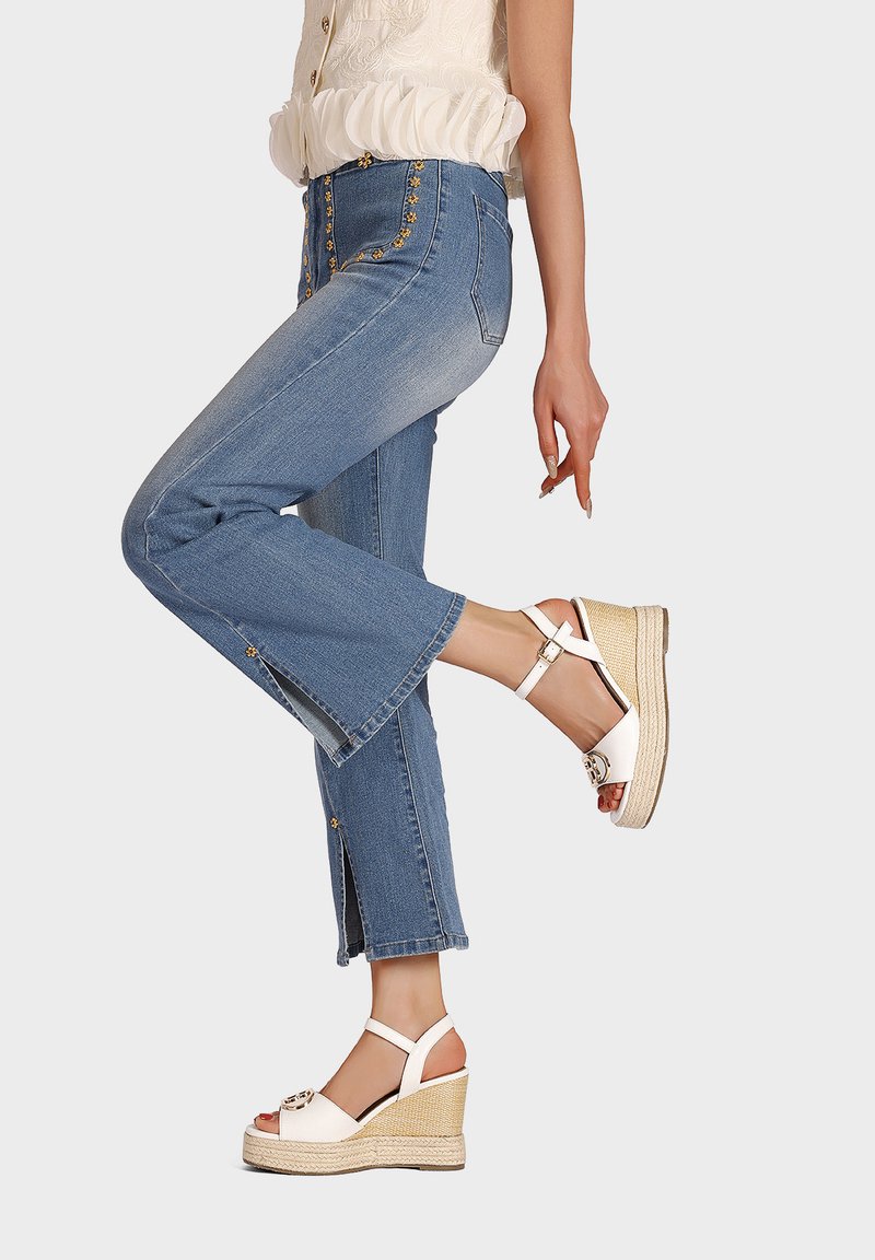 Sandali con tacco a cuneo bianchi con una piattaforma testurizzata e una fascia con fibbia. Indossati con jeans azzurri chiari con dettagli decorativi in oro e orli sfilacciati.