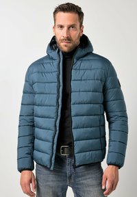 Giacca imbottita isolante color verde smeraldo con motivo trapuntato orizzontale, cappuccio con cappuccio attaccato, cerniera frontale e dettagli neri, abbinata a jeans scuri.