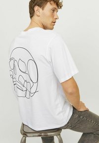 Hvid bomulds T-shirt med en sort stregtegning af et kranium på ryggen. Korte ærmer og en afslappet pasform, kombineret med grå jeans.