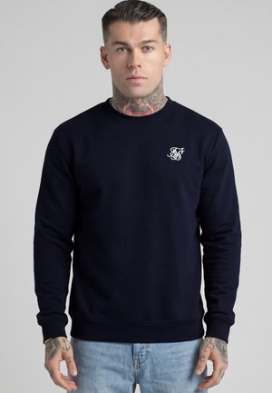 Hombre con tatuajes en el cuello y las manos que lleva una sudadera azul marino con logo blanco en el pecho y vaqueros azul claro, de pie frente a fondo gris.