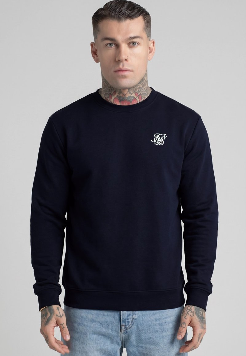 Mann mit Hals- und Handtattoos, der einen dunkelblauen Pullover mit weißem Logo auf der Brust und hellblaue Jeans trägt, steht vor einem grauen Hintergrund.