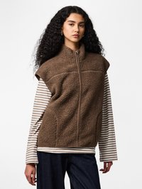 Hosszú, göndör hajú nő, barna cipzáras sherpa mellényt visel bézs és fehér csíkos hosszú ujjú ing fölött, sötét farmernadrággal.