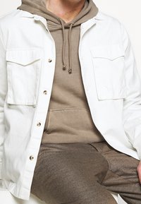 Camicia bianca con bottoni e due tasche sul petto sopra un felpa beige. Tessuto liscio con una vestibilità rilassata e sottili accenti color taupe.