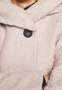 Ljust rosa ullblandad kappa med texturerat tyg, bred krage och en stor svart knapp. Visar en del av armen med ett svart innerskikt.