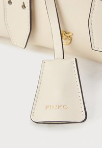 Etiqueta de pele da cor creme com costura escura à volta, destacando o nome da marca "PINKO" em relevo dourado. Ferragens de latão visíveis ao fundo.
