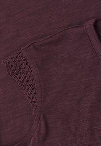 Gros plan d'une manche de chemise en tissu grenat avec un détail en crochet tricoté près de la couture de l'épaule.