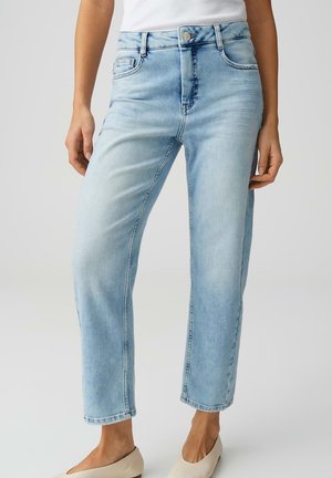 Vrouw met lichtblauwe high-waisted rechte jeans, een wit topje en beige instapschoenen, staand voor een effen achtergrond.