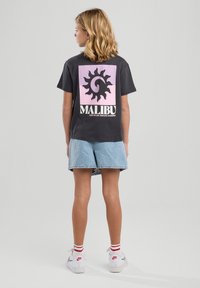 Mustan T-paidan jossa on vaaleanpunainen ja musta auringonmuotoinen design, "MALIBU" -teksti, yhdistettynä farkkushortsien ja valkoisten kenkien kanssa, joissa on punaisia yksityiskohtia.