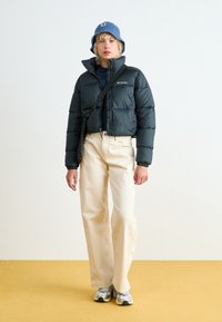 Svart pufferjakke med cropped design og logo, kombinert med vide beige bukser og grå sneakers. Blå strikket bøttehatt fullfører antrekket.