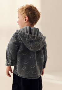 Giacca di denim grigia con cappuccio, caratterizzata da un motivo ripetuto di faccine sorridenti. Maniche lunghe con polsini con bottoni e vestibilità comoda.