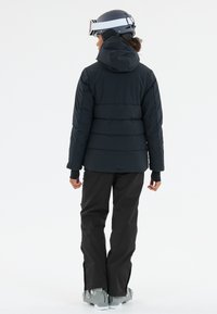 Zwarte pufferjas met capuchon, voorzien van een horizontaal gestikt ontwerp. Gecombineerd met zwarte sneeuwbroeken en skischaatsen. Helm gedragen op het hoofd.