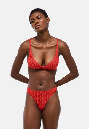 Femme portant un ensemble bikini rouge texturé avec un haut triangle et un bas taille haute, debout sur un fond clair uni, les mains dans le dos.