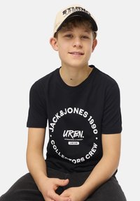 Camiseta de algodón negra con un logotipo gráfico redondo en blanco; combinada con una gorra beige con texto negro. Modelo sentado con las manos en las rodillas.