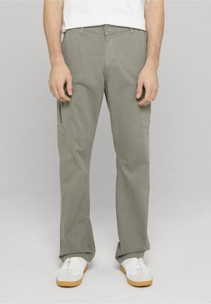 Homme portant un pantalon cargo gris clair avec des poches latérales et des baskets blanches avec semelles en gomme, debout devant un fond uni.