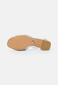 Sølv hæl sandaler med en glat overdel, minimalistisk design og en tan gummisåle med teksturert grep og sirkulært logo-detall.