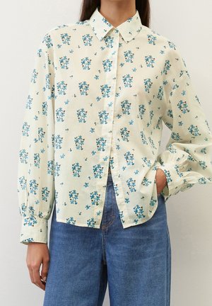 Vrouw draagt een crèmekleurig overhemd met blauw bloemenprint en pofmouwen, gecombineerd met een hoge taille blauwe spijkerbroek, één hand in de zak.