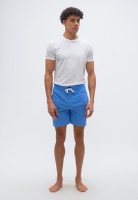 Νεαρός άνδρας στέκεται ξυπόλητος, φοράει λευκό κοντομάνικο πουκάμισο και μπλε σορτς με κορδόνι, με φόντο έναν απλό λευκό τοίχο.