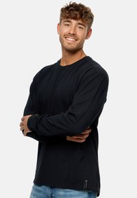 Schwarzes Langarmshirt aus strukturiertem Stoff, mit Rundhalsausschnitt und seitlichem Etikett, kombiniert mit hellblauen Jeans.