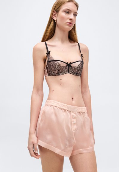 Γυναίκα που φοράει μαύρο δαντελωτό σουτιέν με φιόγκους και διάφανο ροζ σατέν σορτς, στέκεται μπροστά σε απλό φωτεινό φόντο.