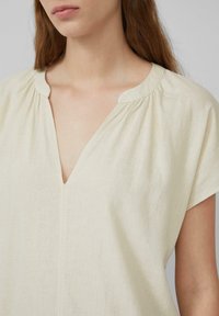 Hellbeige, kurzärmliges Oberteil mit V-Ausschnitt, das an den Schultern dezent gerafft ist und aus einem strukturierten Stoff gefertigt ist.