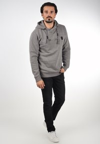 Sudadera gris con cordones y un pequeño logo, combinada con pantalones negros y zapatillas blancas. Tela suave, diseño casual.
