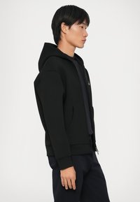 Svart hettejakke med glidelås foran, sidelommer og ribbestrikkede mansjetter, laget av et mykt stoff, vist på en modell i profil.