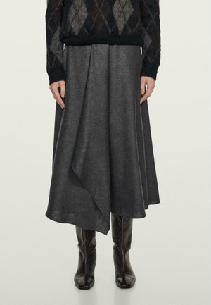 Femme portant un pull à losanges sombre, une jupe midi asymétrique gris charbon et des bottes en cuir noires montant jusqu'aux genoux, debout devant un fond uni.