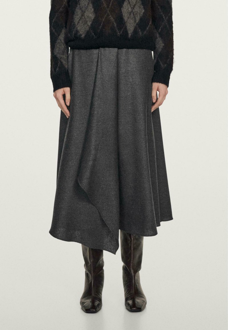 Femme portant un pull à losanges sombre, une jupe midi asymétrique gris charbon et des bottes en cuir noires montant jusqu'aux genoux, debout devant un fond uni.