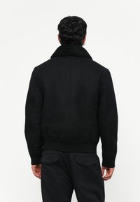 Chaqueta bomber negra con cuello de piel de oveja, puños y dobladillo acanalados. La parte trasera tiene un acabado liso y está diseñada para un ajuste ceñido.