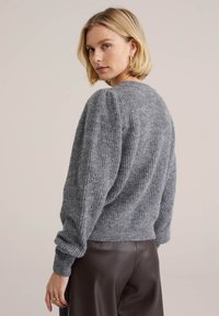 Gråstickad tröja med en strukturerad yta, rund halsringning och lätt puffade ärmar, matchad med mörkbruna läderbyxor.