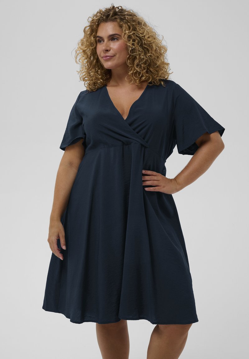 Femme aux cheveux bouclés portant une robe portefeuille bleu marine mi-longue avec des manches courtes volantées, debout avec une main sur la hanche.