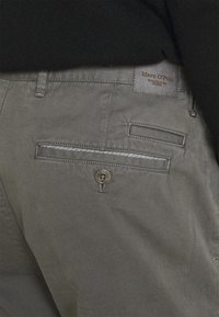 Zbliżenie na tylnią kieszeń khaki spodni z zapięciem na guzik oraz wszytą metką marki "Marc O'Polo" na pasku.