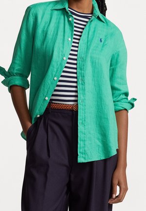 Henkilö pukeutuneena käärityllä vihreällä pitkähihhaisella paidalla, jonka alla on laivastonsininen ja valkoraidallinen paita, ruskea punottu vyö ja tummat väljät housut.