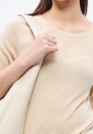 Femme portant un haut en maille beige tenant la bandoulière d’un sac à bandoulière crème avec la main gauche sur fond blanc.