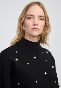 Maglione nero a maglia con collo alto, impreziosito da accenti metallici rotondi disposti su tutta la parte anteriore in un motivo sparso.
